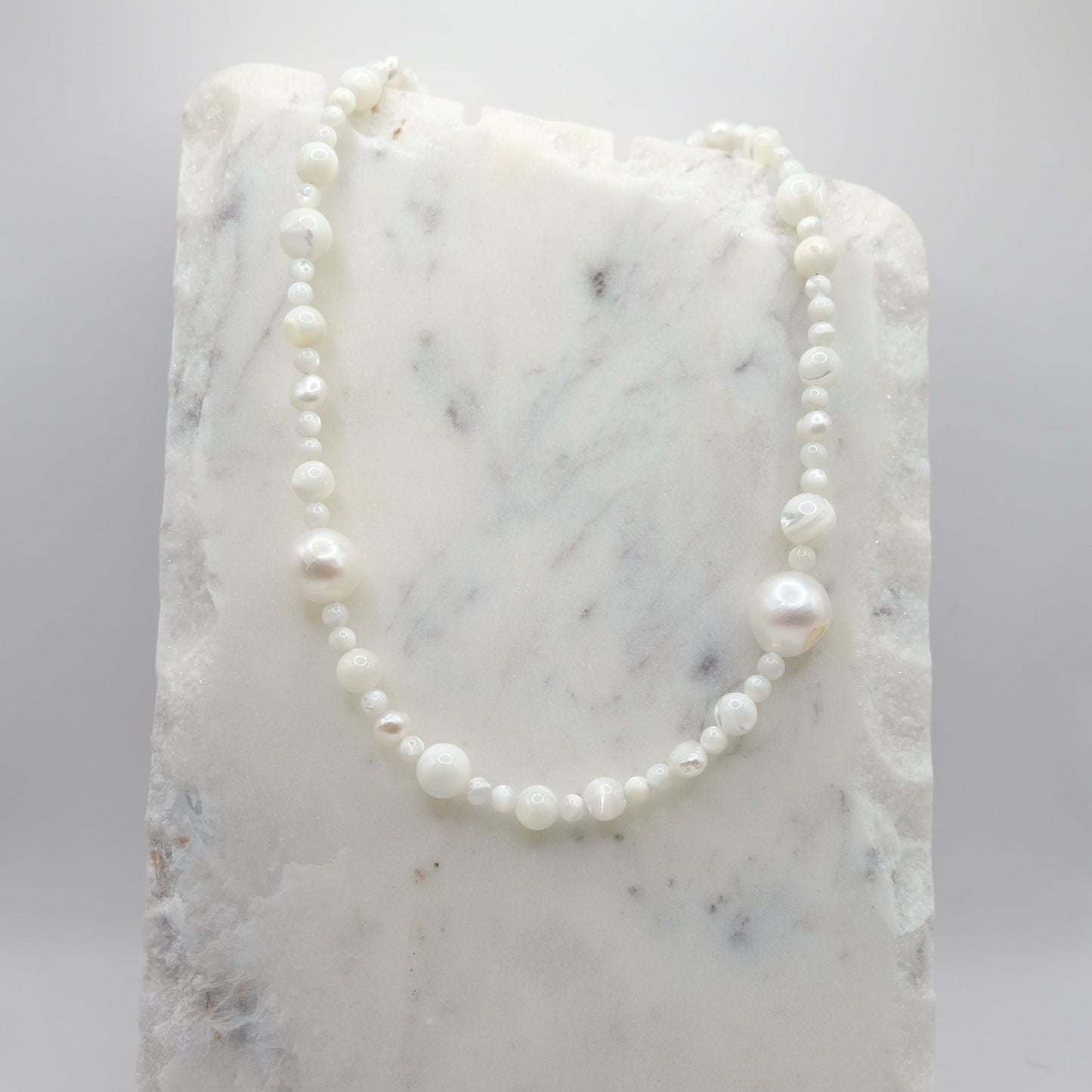 A beaded necklace composed of mother of pearl beads in mixed sizes - featuring scattered freshwater pearl nuggets, shown on a white marble background.