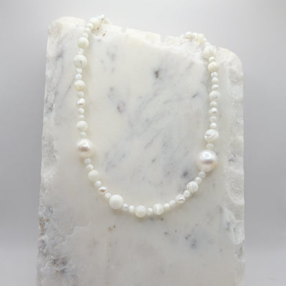 A beaded necklace composed of mother of pearl beads in mixed sizes - featuring scattered freshwater pearl nuggets, shown on a white marble background.
