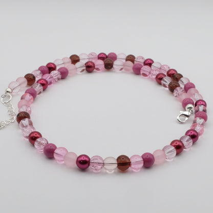 A handmade beaded necklace featuring soft pink, berry and clear glass beads arranged in a playful pattern, displayed on a white marble jewellery stand.