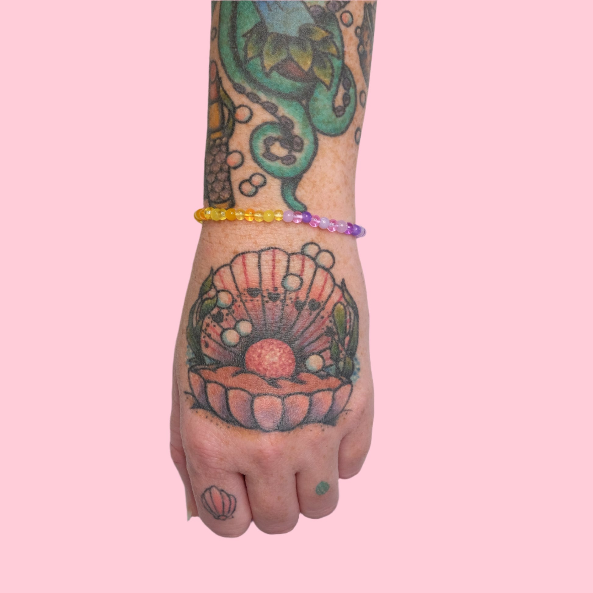 A tattooed arm featuring an octopus and seashell design wears an elastic beaded bracelet of purple and yellow beads, displayed against a solid pink background.