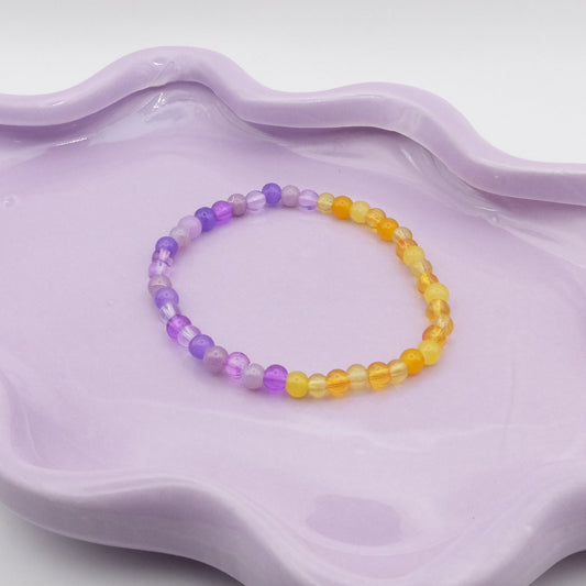 An elastic beaded bracelet made from 4mm glass beads, featuring two halves—one side a mix of purple beads and and the other side a mix of yellow beads, resting on a wavy lavender ceramic dish against a light background.
