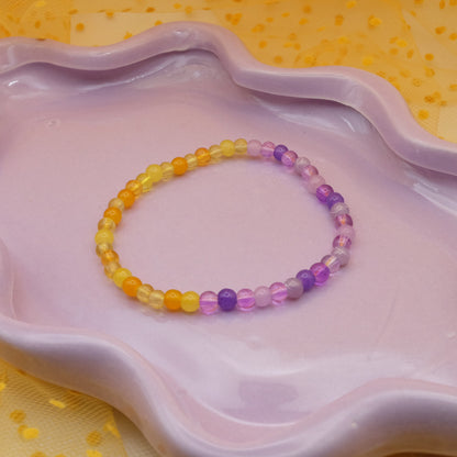 An elastic beaded bracelet made from 4mm glass beads, featuring two halves—one side a mix of purple beads and and the other side a mix of yellow beads, displayed on a wavy lavender ceramic dish with a yellow polka-dot tulle backdrop.