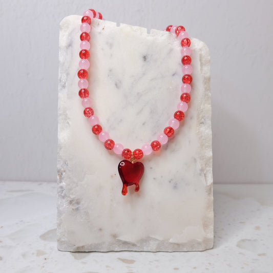 Necklace with red and pink beads and a heart-shaped pendant on a marble stand.