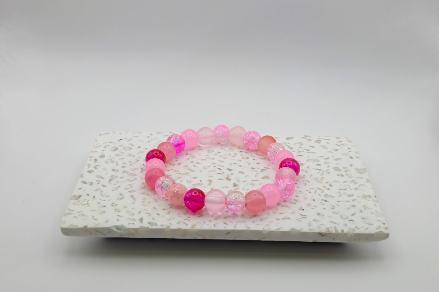 An elastic beaded bracelet composed of 8mm pink glass beads - featuring frosted, crackle, and solid textures, shown on a terrazzo tile in a light studio setting.