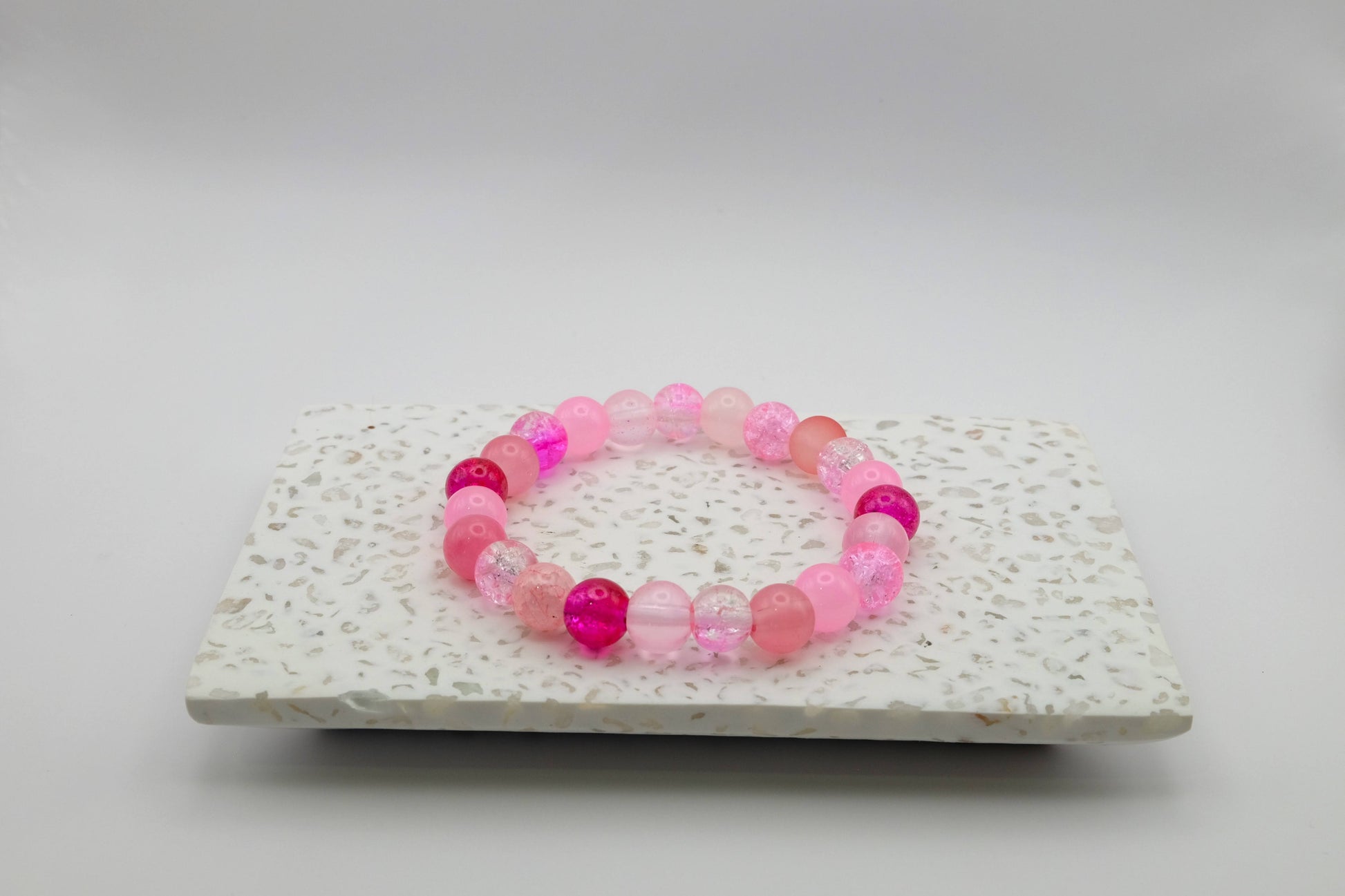 An elastic beaded bracelet composed of 8mm pink glass beads - featuring frosted, crackle, and solid textures, shown on a terrazzo tile in a light studio setting.