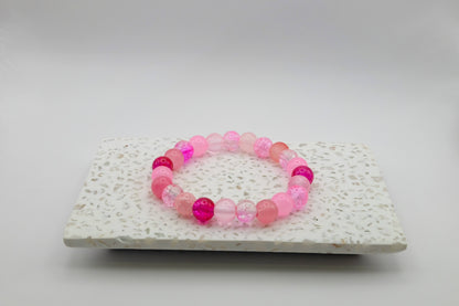 An elastic beaded bracelet composed of 8mm pink glass beads - featuring frosted, crackle, and solid textures, shown on a terrazzo tile in a light studio setting.