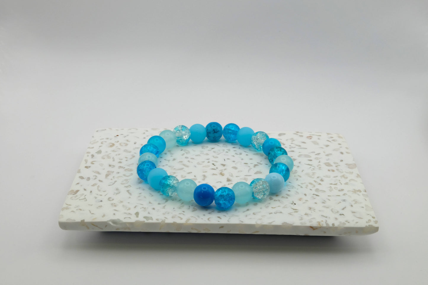 An elastic beaded bracelet composed of 8mm blue glass beads in a mix of blues - featuring frosted, crackle, and solid textures, on a terrazzo tile.