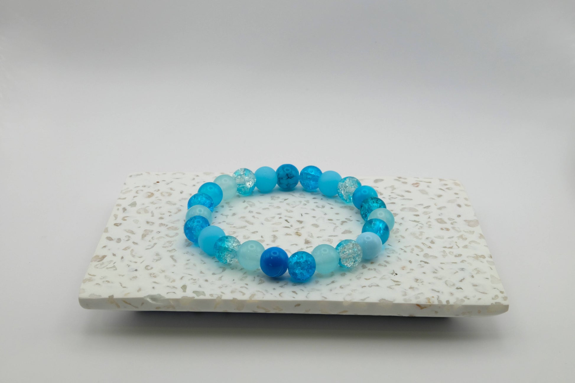 An elastic beaded bracelet composed of 8mm blue glass beads in a mix of blues - featuring frosted, crackle, and solid textures, on a terrazzo tile.