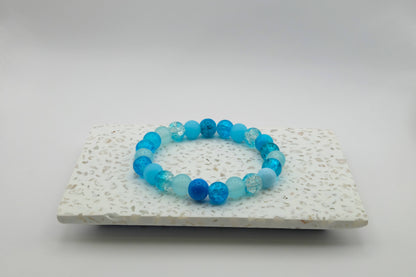An elastic beaded bracelet composed of 8mm blue glass beads in a mix of blues - featuring frosted, crackle, and solid textures, on a terrazzo tile.