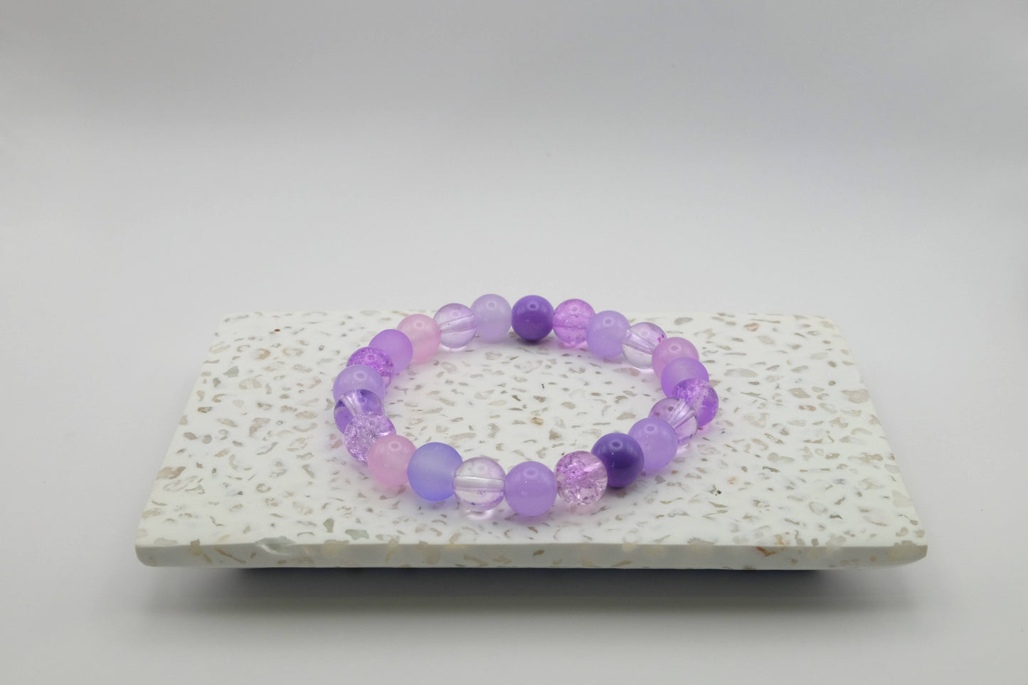 An elastic beaded bracelet composed of 8mm purple glass beads featuring frosted, crackle, and solid finishes, shown on a terrazzo tile background. 