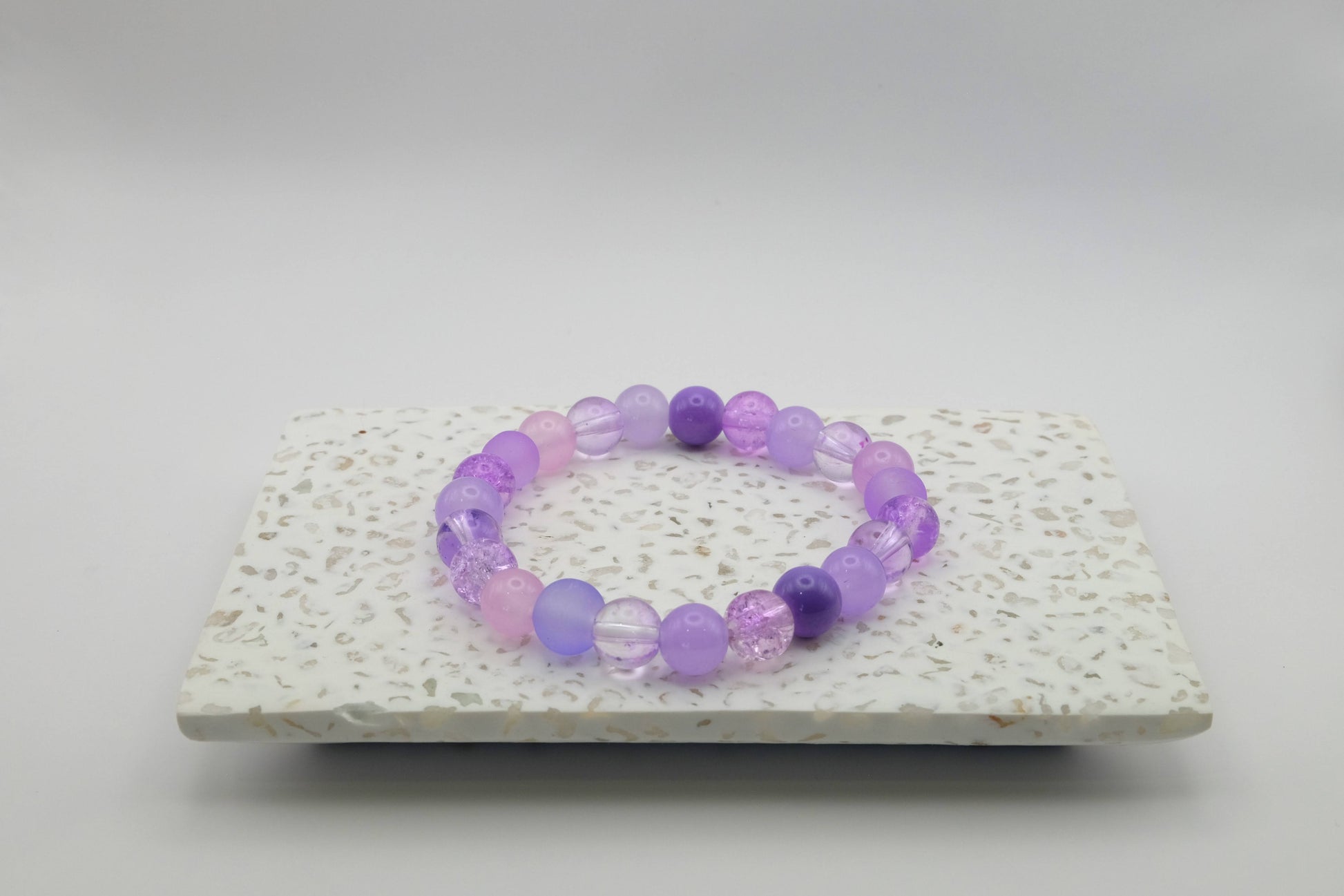 An elastic beaded bracelet composed of 8mm purple glass beads featuring frosted, crackle, and solid finishes, shown on a terrazzo tile background. 