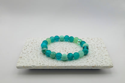 An elastic beaded bracelet composed of 8mm aqua, teal and mint green glass beads - featuring crackle, frosted, and glossy finishes, shown on a terrazzo tile background.