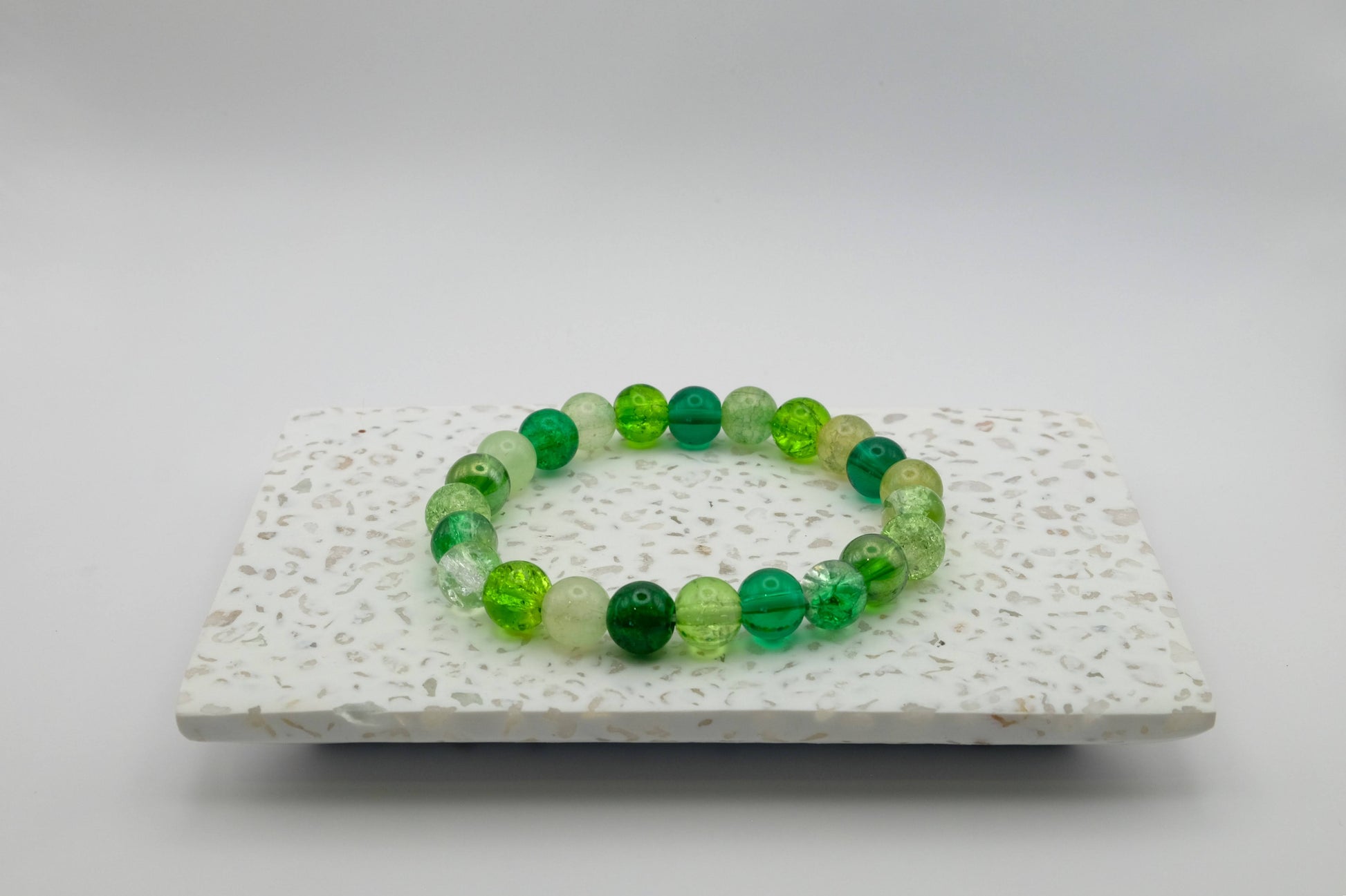 An elastic beaded bracelet composed of 8mm green glass beads - from soft sage to emerald - featuring frosted and crackle textures, shown on a terrazzo tile base.