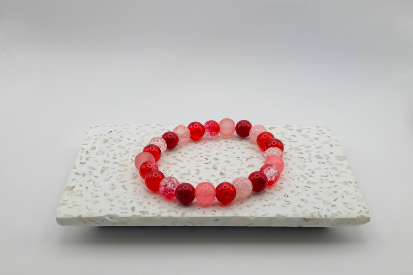 An elastic beaded bracelet composed of 8mm glass beads in different shades of red—featuring crackle, frosted, and glossy textures, displayed on a terrazzo tile.