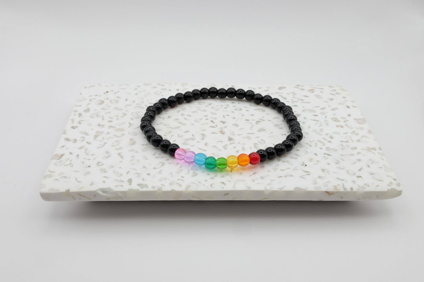 An elastic beaded bracelet composed of 4mm black glass beads accented by eight vibrant beads, one each in red, orange, yellow, green, aqua, blue, purple and pink shown on a terrazzo tile in front of a minimalist light backdrop.