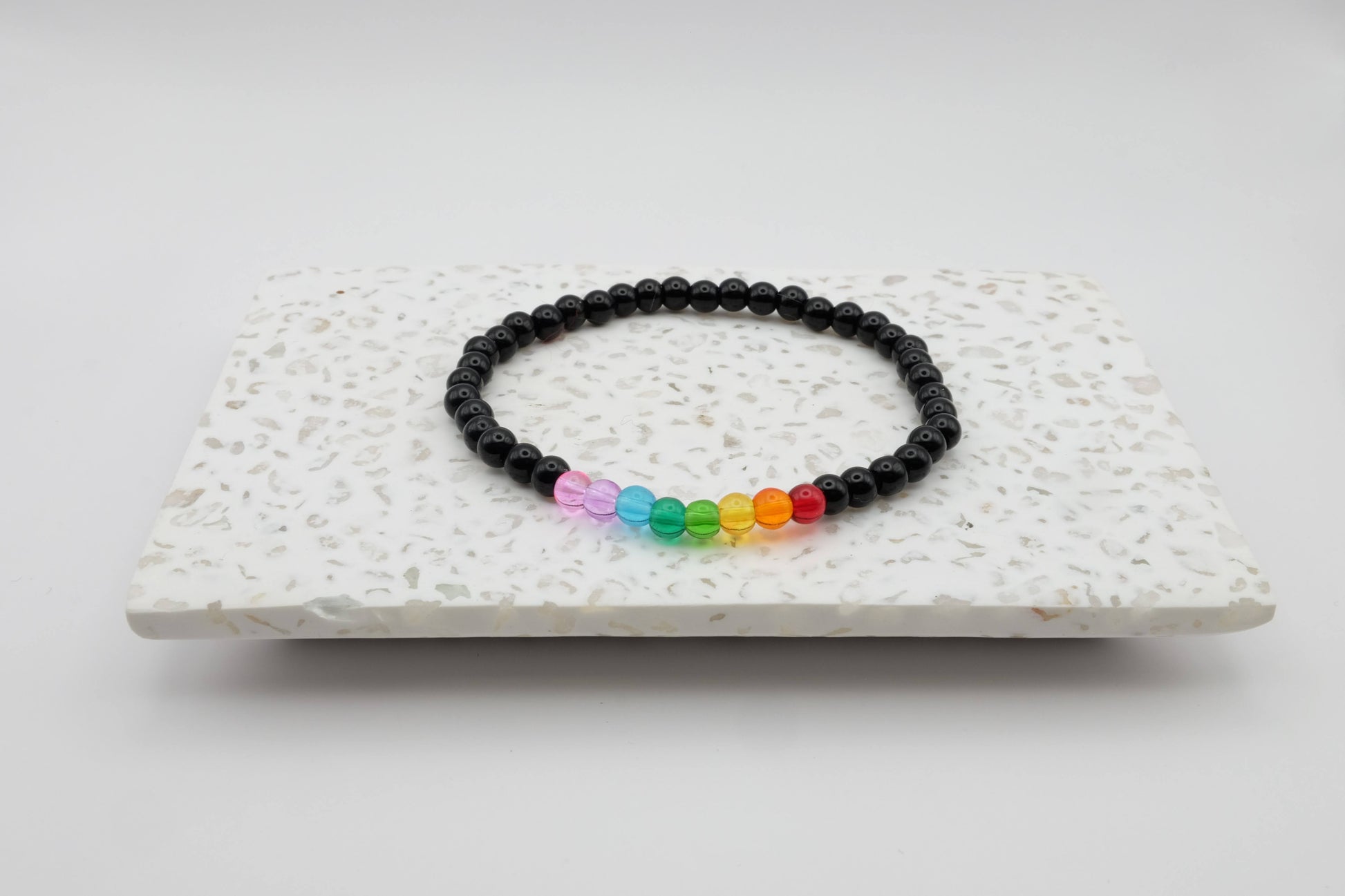 An elastic beaded bracelet composed of 4mm black glass beads accented by eight vibrant beads, one each in red, orange, yellow, green, aqua, blue, purple and pink shown on a terrazzo tile in front of a minimalist light backdrop.