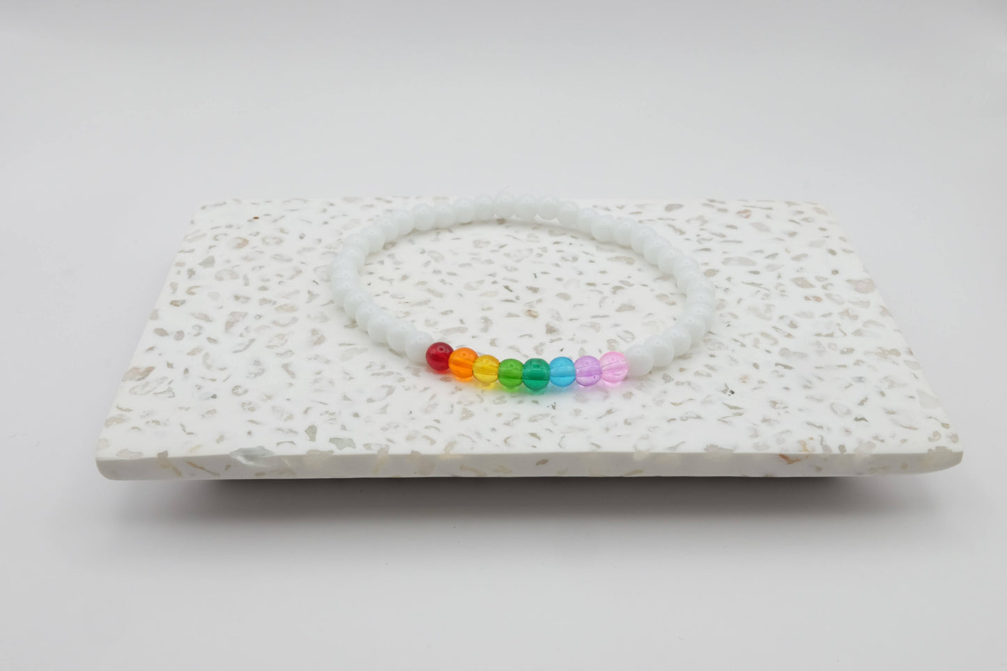 An elastic beaded bracelet composed of 4mm white glass beads accented by eight vibrant beads, one each in red, orange, yellow, green, aqua, blue, purple and pink shown on a terrazzo tile in front of a minimalist light backdrop.