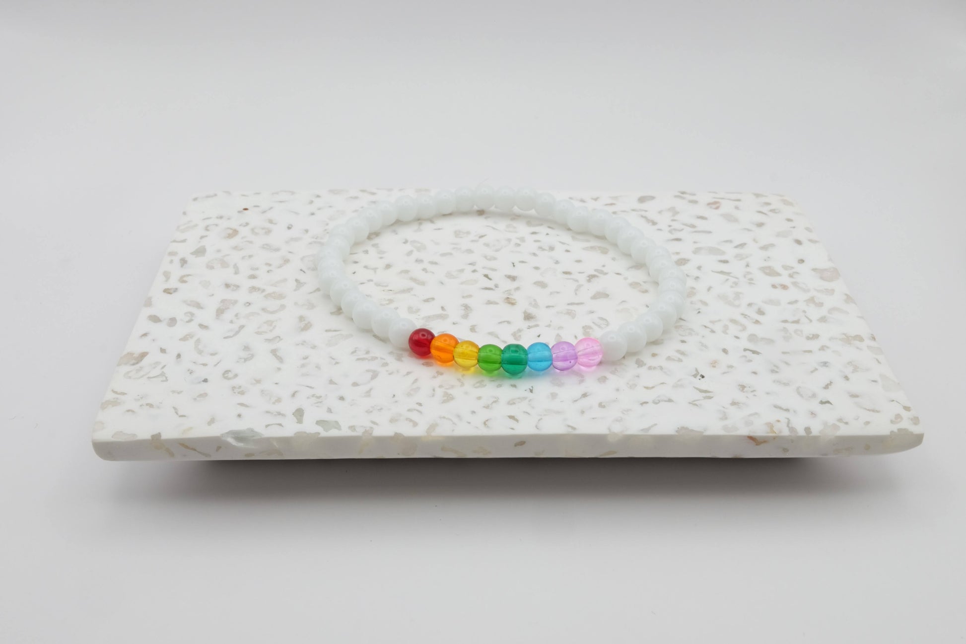An elastic beaded bracelet composed of 4mm white glass beads accented by eight vibrant beads, one each in red, orange, yellow, green, aqua, blue, purple and pink shown on a terrazzo tile in front of a minimalist light backdrop.
