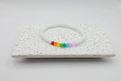 An elastic beaded bracelet composed of 4mm white glass beads accented by eight vibrant beads, one each in red, orange, yellow, green, aqua, blue, purple and pink shown on a terrazzo tile in front of a minimalist light backdrop.