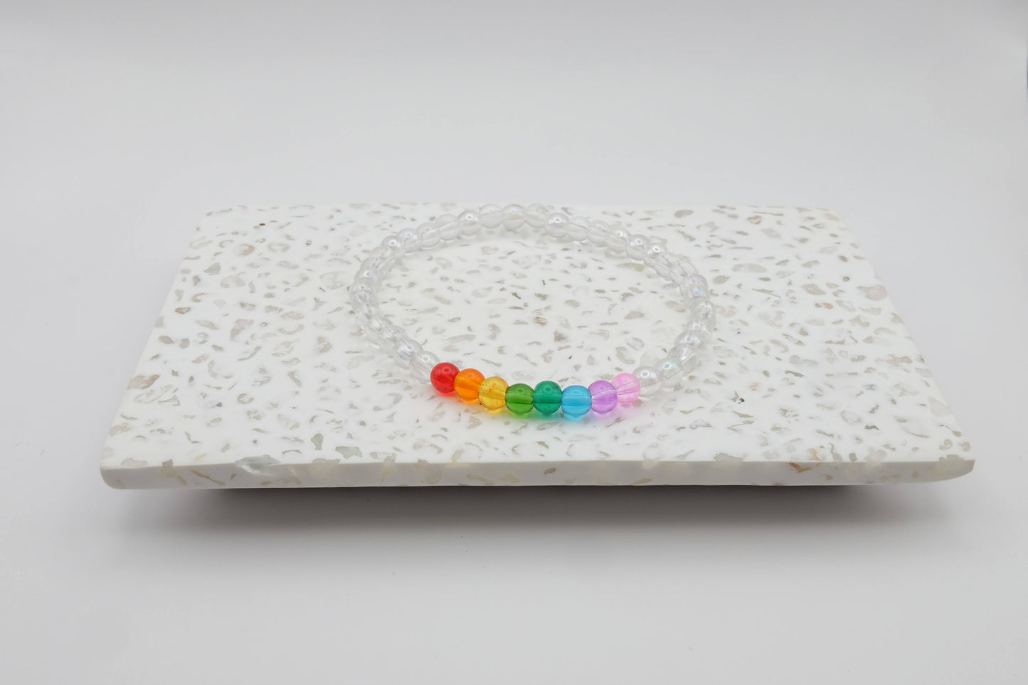 An elastic beaded bracelet composed of 4mm clear glass beads accented by eight vibrant beads, one each in red, orange, yellow, green, aqua, blue, purple and pink shown on a terrazzo tile in front of a minimalist light backdrop.