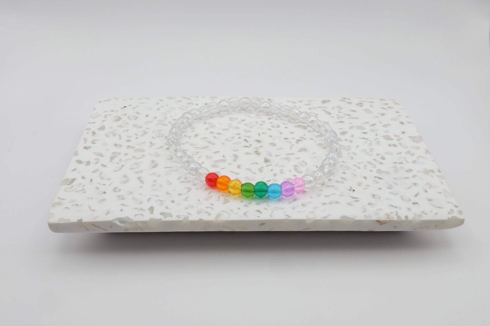 An elastic beaded bracelet composed of 4mm clear glass beads accented by eight vibrant beads, one each in red, orange, yellow, green, aqua, blue, purple and pink shown on a terrazzo tile in front of a minimalist light backdrop.