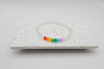 An elastic beaded bracelet composed of 4mm clear glass beads accented by eight vibrant beads, one each in red, orange, yellow, green, aqua, blue, purple and pink shown on a terrazzo tile in front of a minimalist light backdrop.