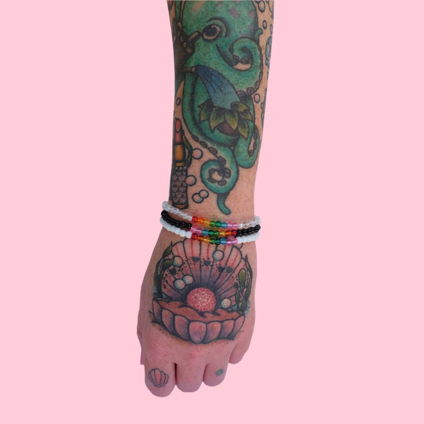 A tattooed arm wearing a stack of three elastic beaded bracelets made with 4mm glass beads, one in black, white, and clear and each accented by 8 vibrant beads rainbow tones. 