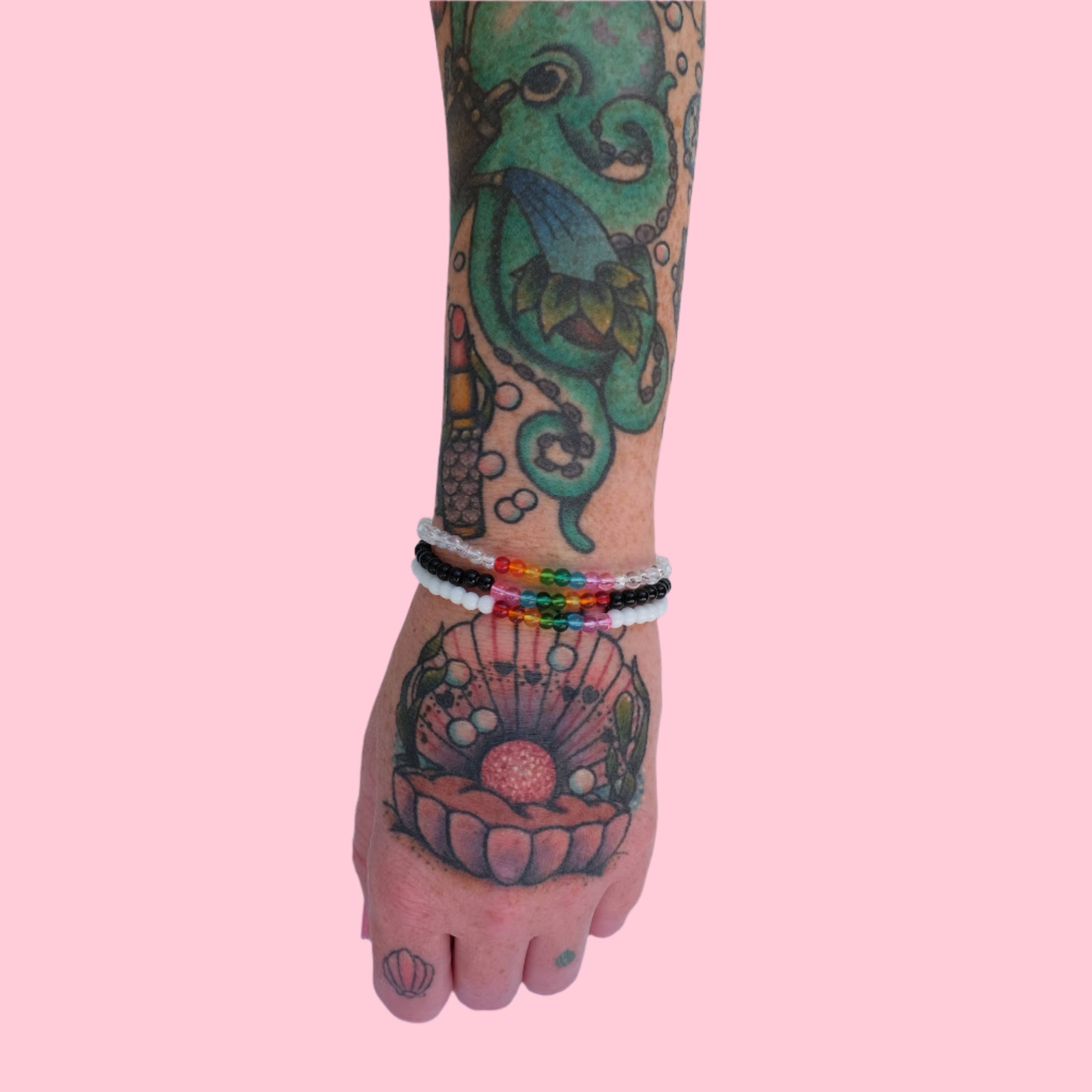 A tattooed arm wearing a stack of three elastic beaded bracelets made with 4mm glass beads, one in black, white, and clear and each accented by 8 vibrant beads rainbow tones. 