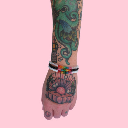 A tattooed arm wearing a stack of three elastic beaded bracelets made with 4mm glass beads, one in black, white, and clear and each accented by 8 vibrant beads rainbow tones. 