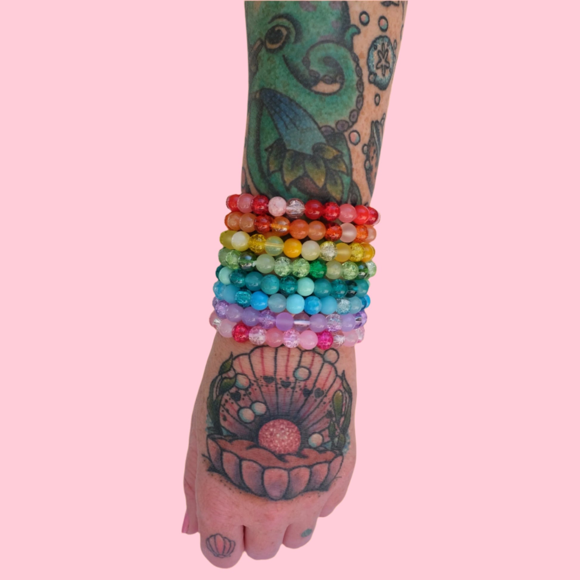 A full rainbow stack of 8 elastic beaded bracelets composed of 8mm glass beads - featuring frosted, crackle, and solid textures in red, orange, yellow, green, aqua, blue, purple, and pink hues. Shown on a tattooed arm.