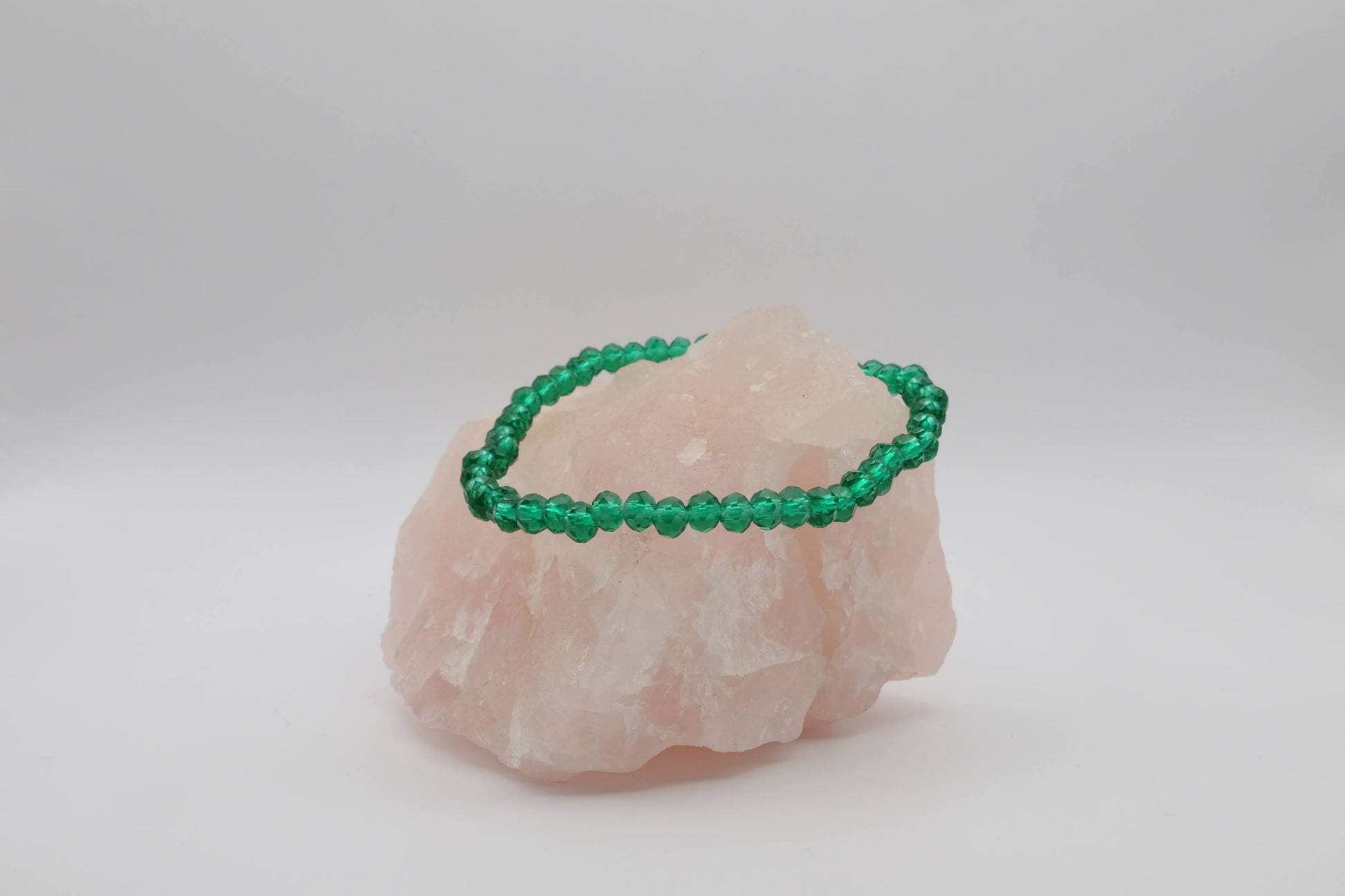 A green beaded bracelet made with faceted beads, elegantly displayed on a light pink crystal stone against a plain white background