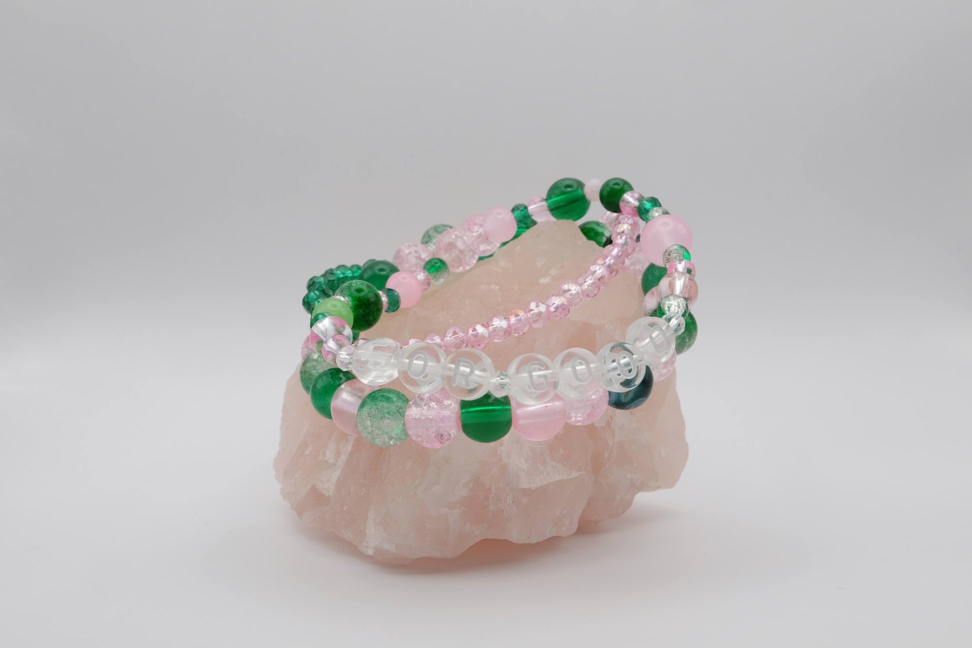 A stack of three bracelets in a mix of green and pink hues and a variety of bead types, placed on a soft pink crystal against a plain white background. One of the bracelets has the words "For Good" spelled out in letter beads.