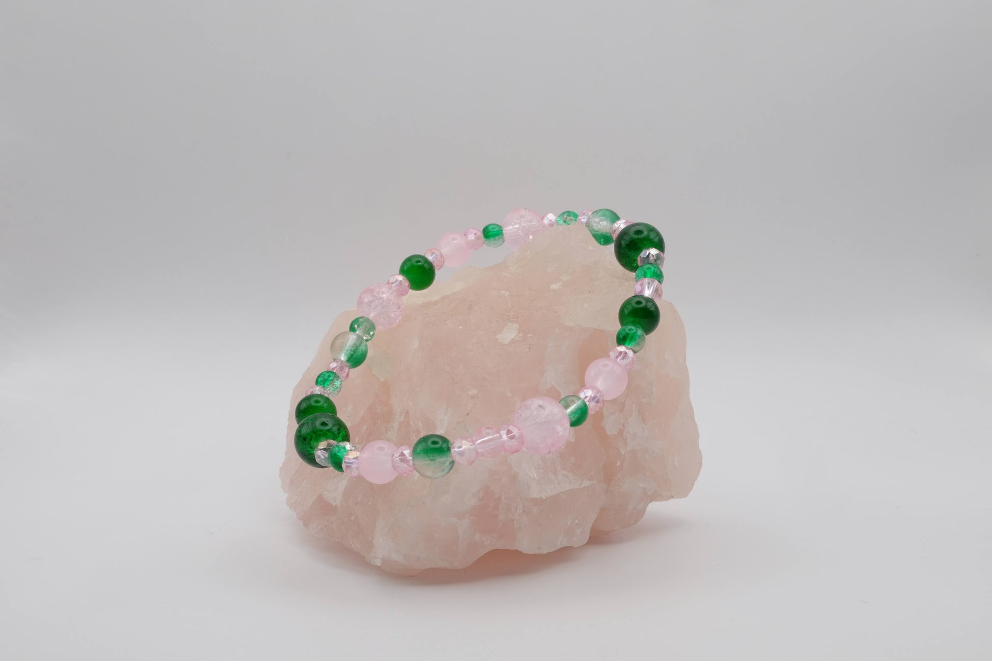 A delicate bracelet featuring a mix of green and pink beads, displayed on a light pink crystal stone against a plain white background.
