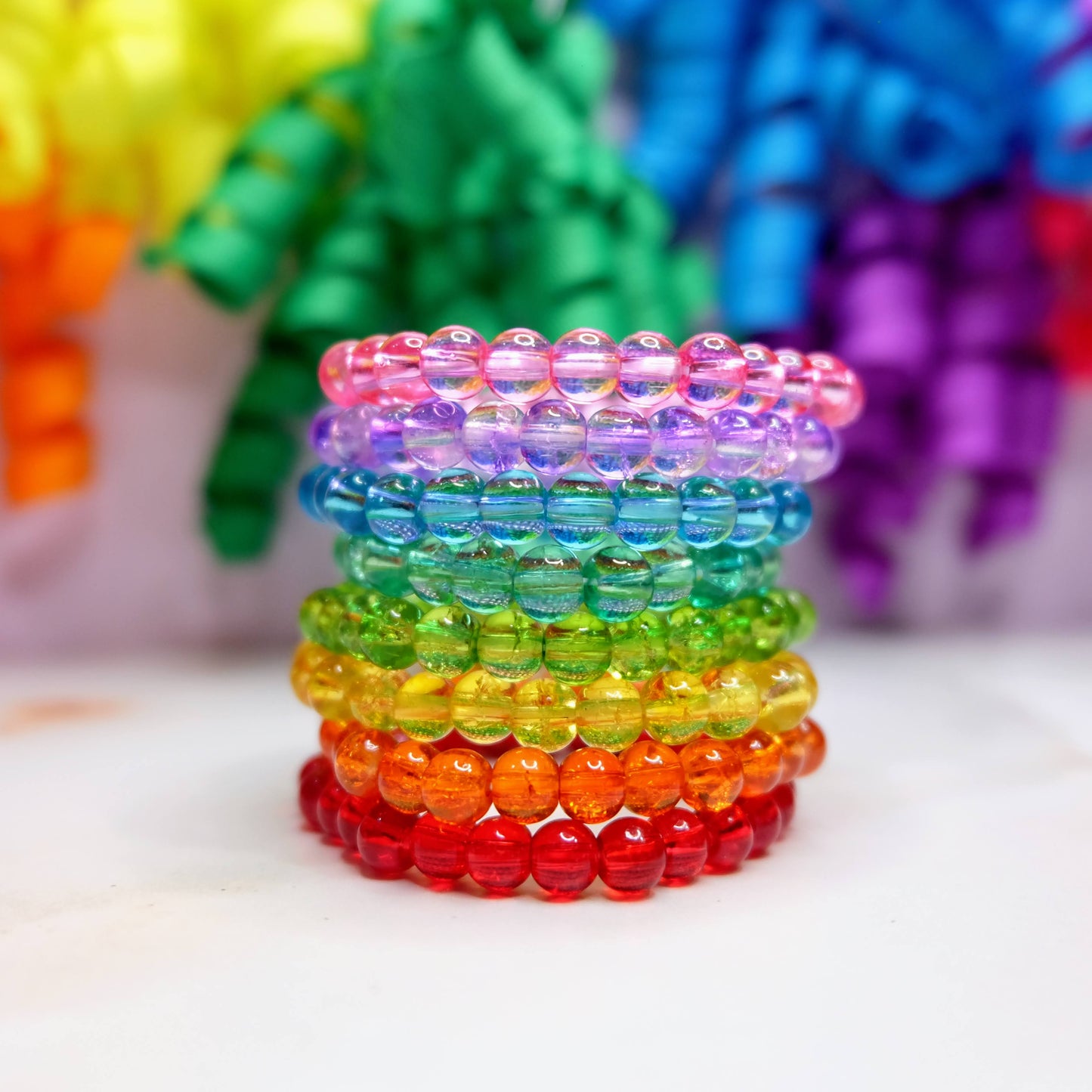 A stack of 8 bracelet, each in a colour of the rainbow - red,  orange, yellow, green,  aqua, blue, purple and pink. Bracelets are stacked up on top of each other and sit in front of a rainbow ribbon background.
