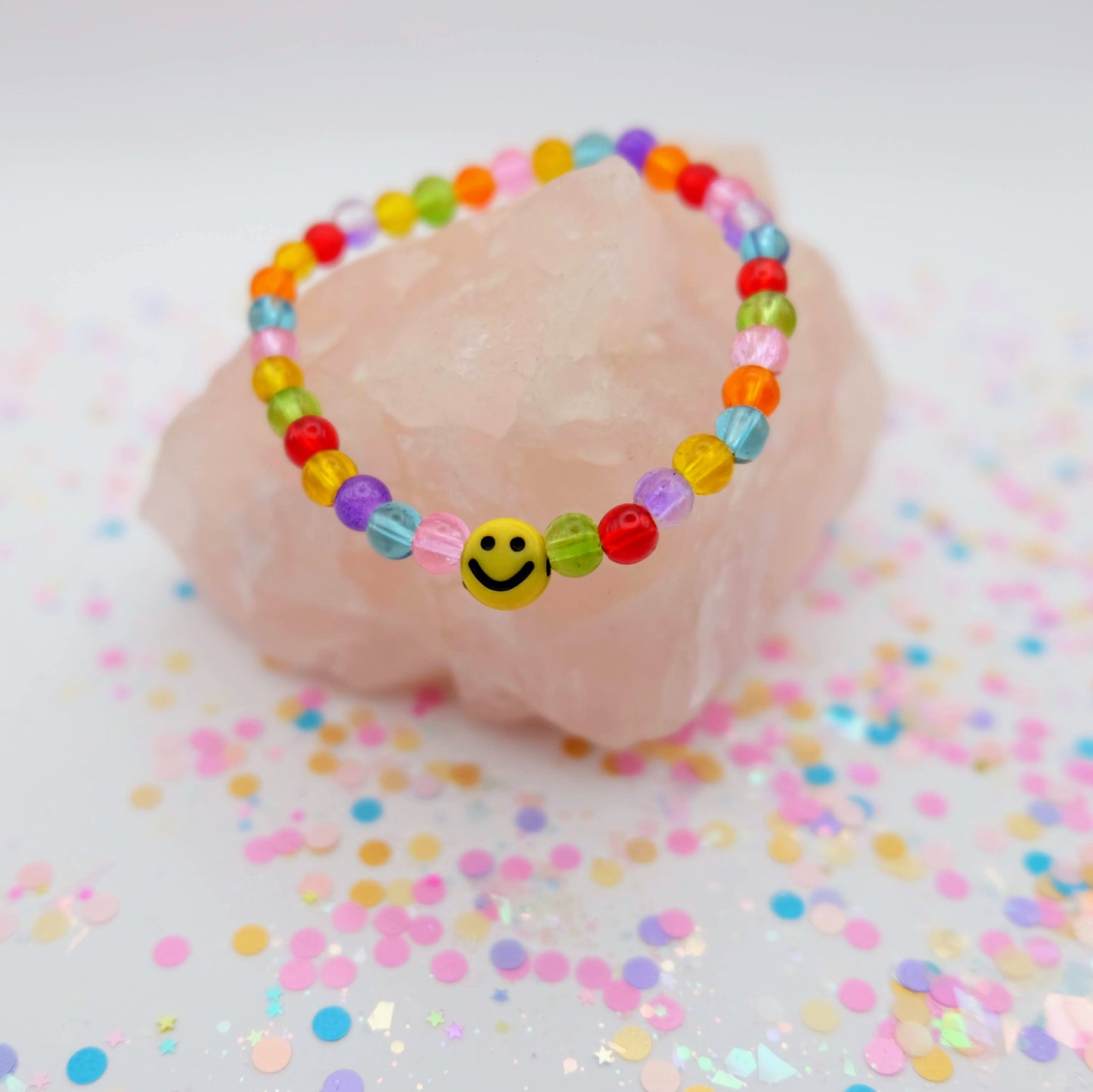 Rainbow beaded bracelet with a smiley face charm on a pink shell with confetti background