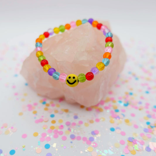 Rainbow beaded bracelet with a smiley face charm on a pink shell with confetti background
