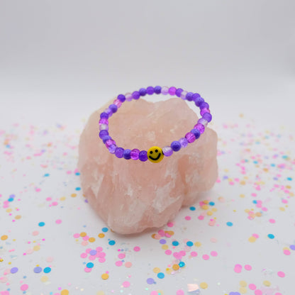 Purple beaded bracelet with a smiley face charm on a pink stone with colorful confetti