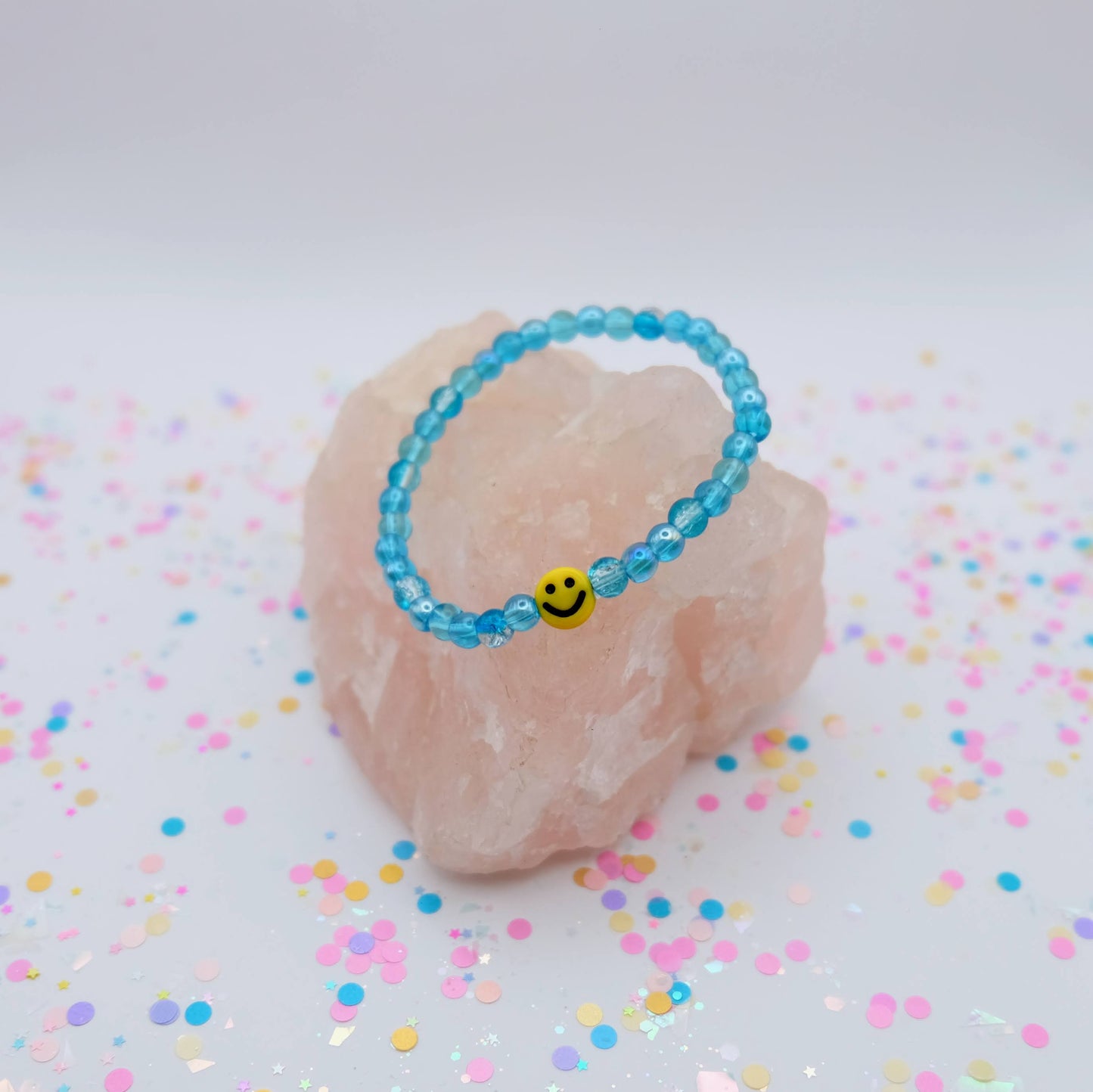 Blue beaded bracelet with a smiley face charm on a pink stone with colorful confetti background
