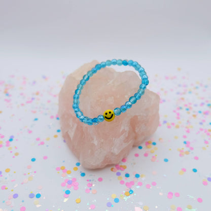 Blue beaded bracelet with a smiley face charm on a pink stone with colorful confetti background
