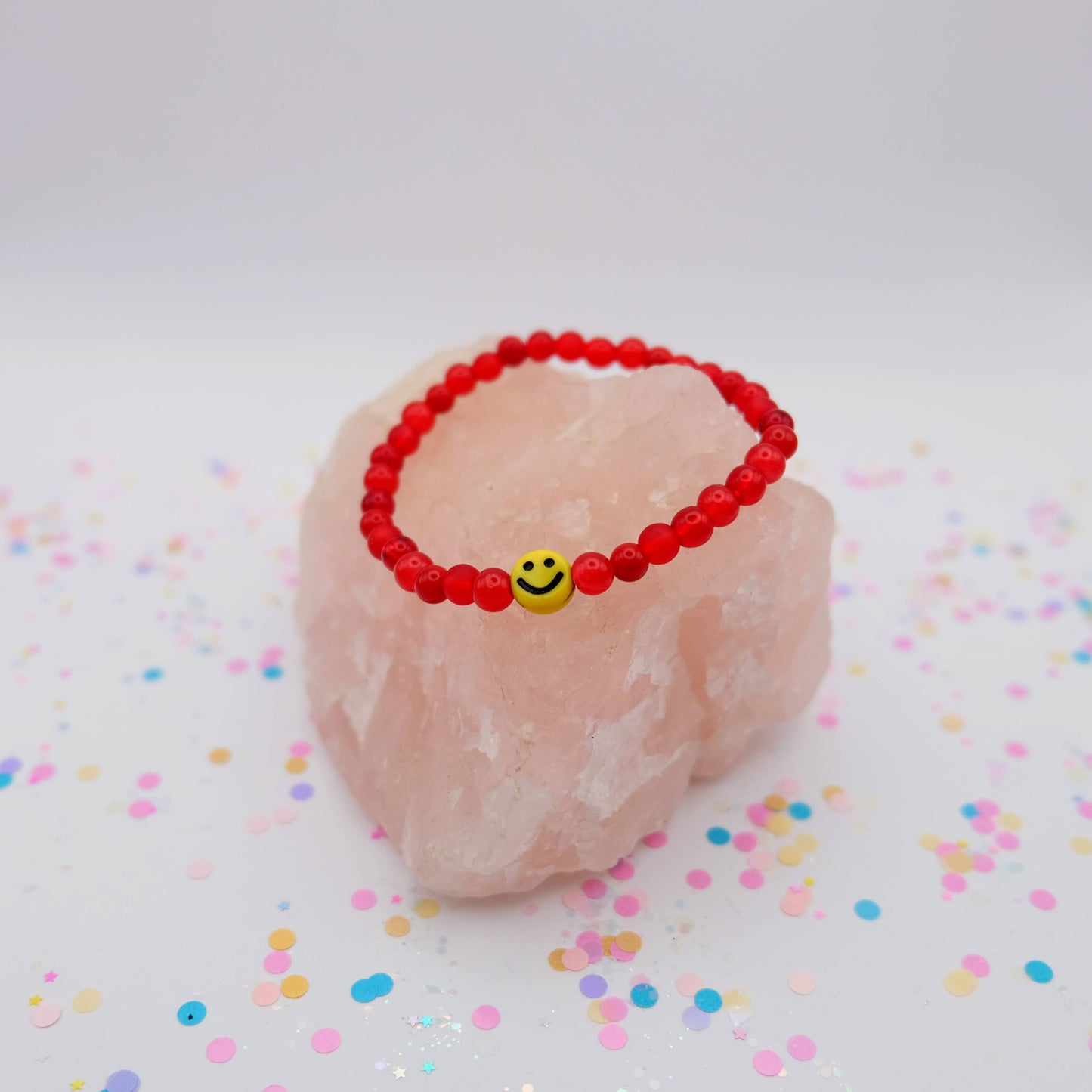 Red beaded bracelet with a smiley face charm on a pink stone with colorful confetti