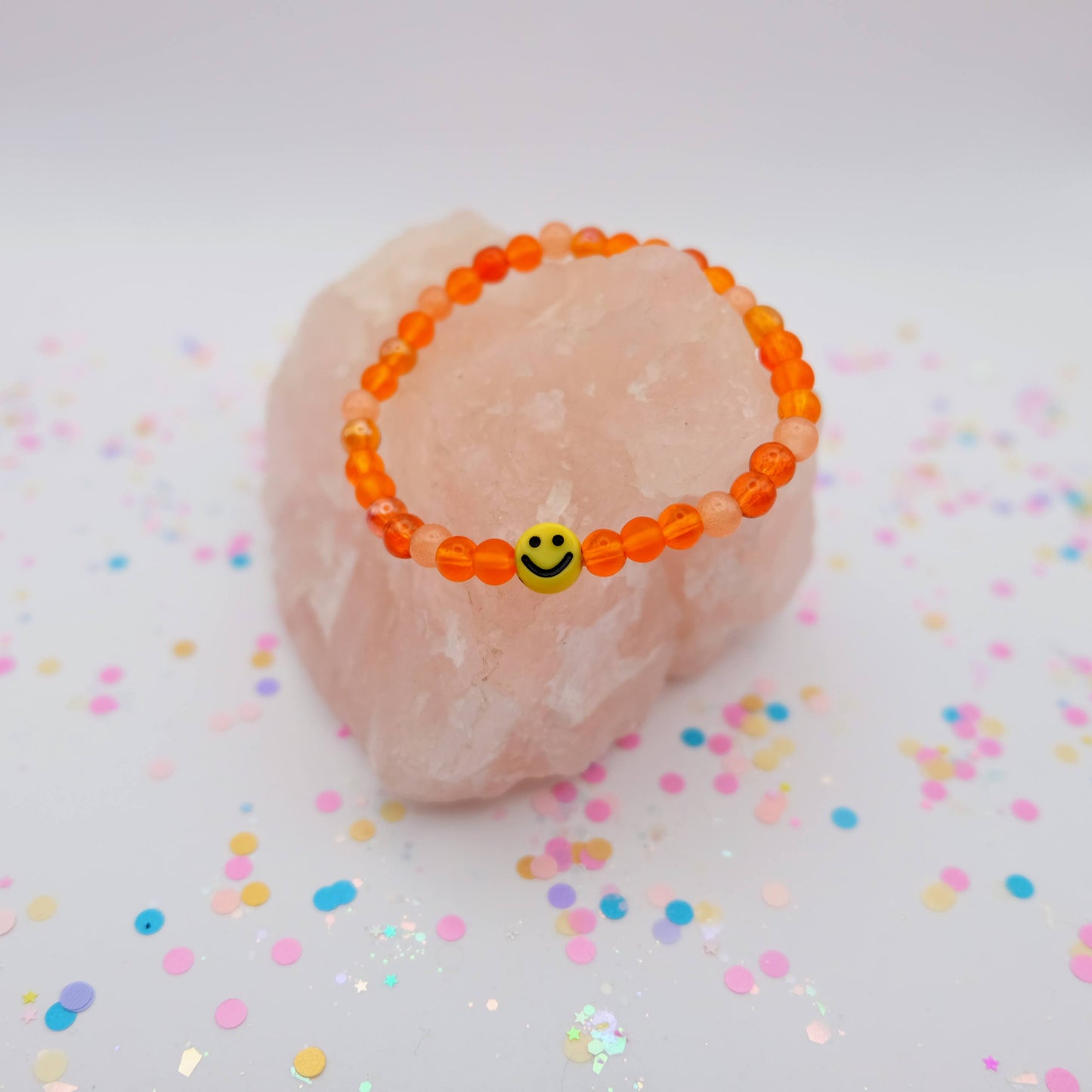 Orange beaded bracelet with a smiley face charm on a pink stone with colorful confetti background