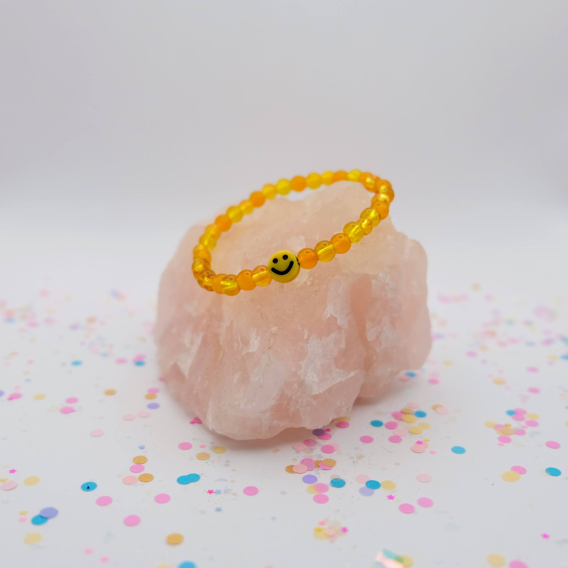 Yellow beaded bracelet with a smiley face charm on a pink stone with colorful confetti