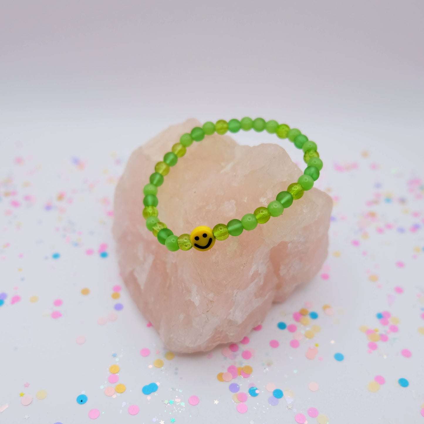 Green beaded bracelet with a smiley face charm on a pink stone with colorful confetti background