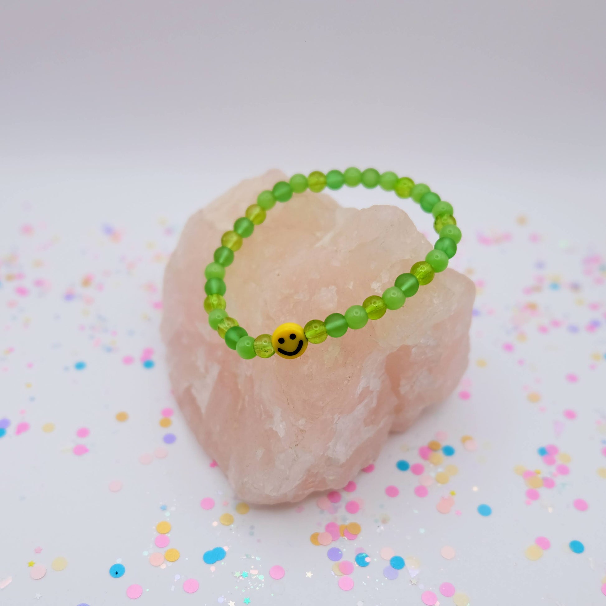 Green beaded bracelet with a smiley face charm on a pink stone with colorful confetti background