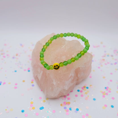 Green beaded bracelet with a smiley face charm on a pink stone with colorful confetti background