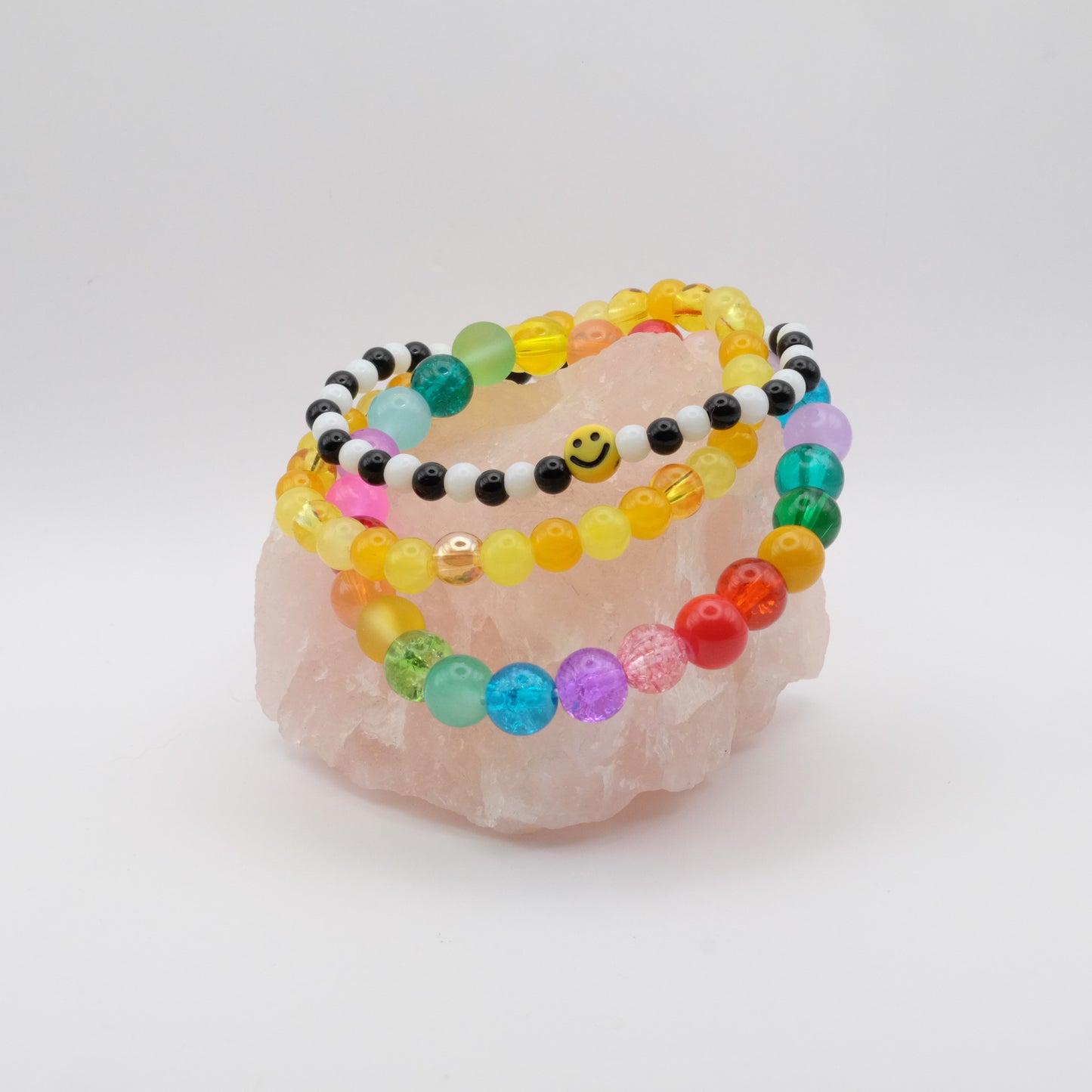 A stack of colourful bracelets, one with a smiley face bead, in the colours of the rainbow on a rose quartz rock