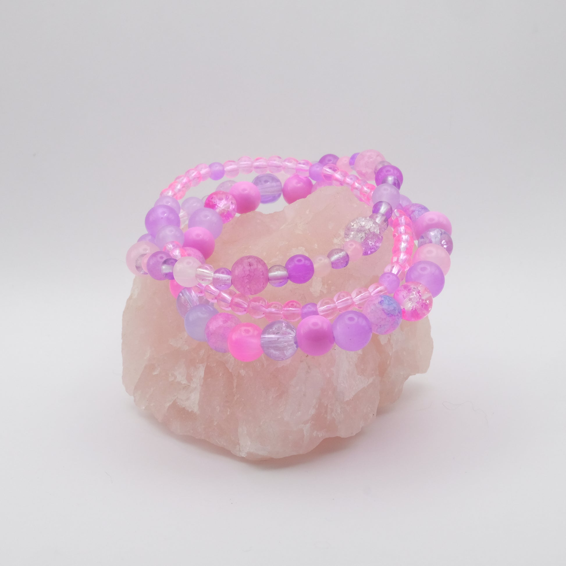 Three beaded bracelets in pink, purple, and clear on a rose quartz stone with a white background
