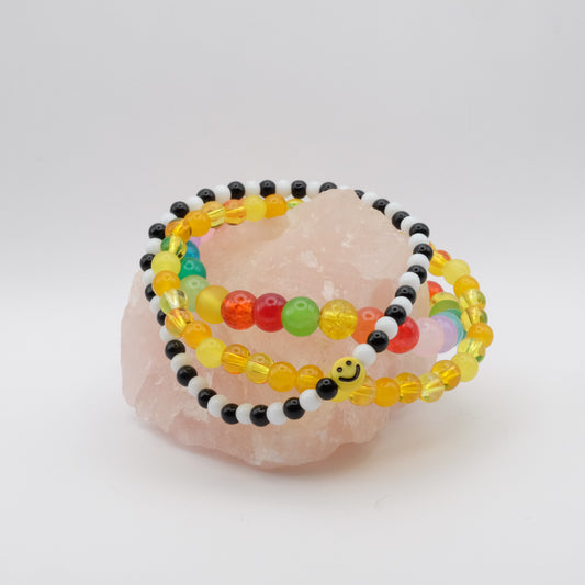 A stack of three elastic beaded bracelets in the colours of the rainbow , one with a smiley face bead, sitting on a rose quartz rock.