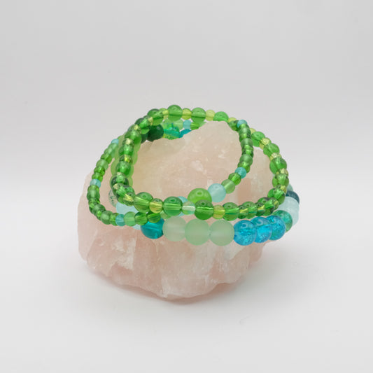 A stack of three elastic beaded bracelets in greens, teals and blues sitting on a rose quartz rock.