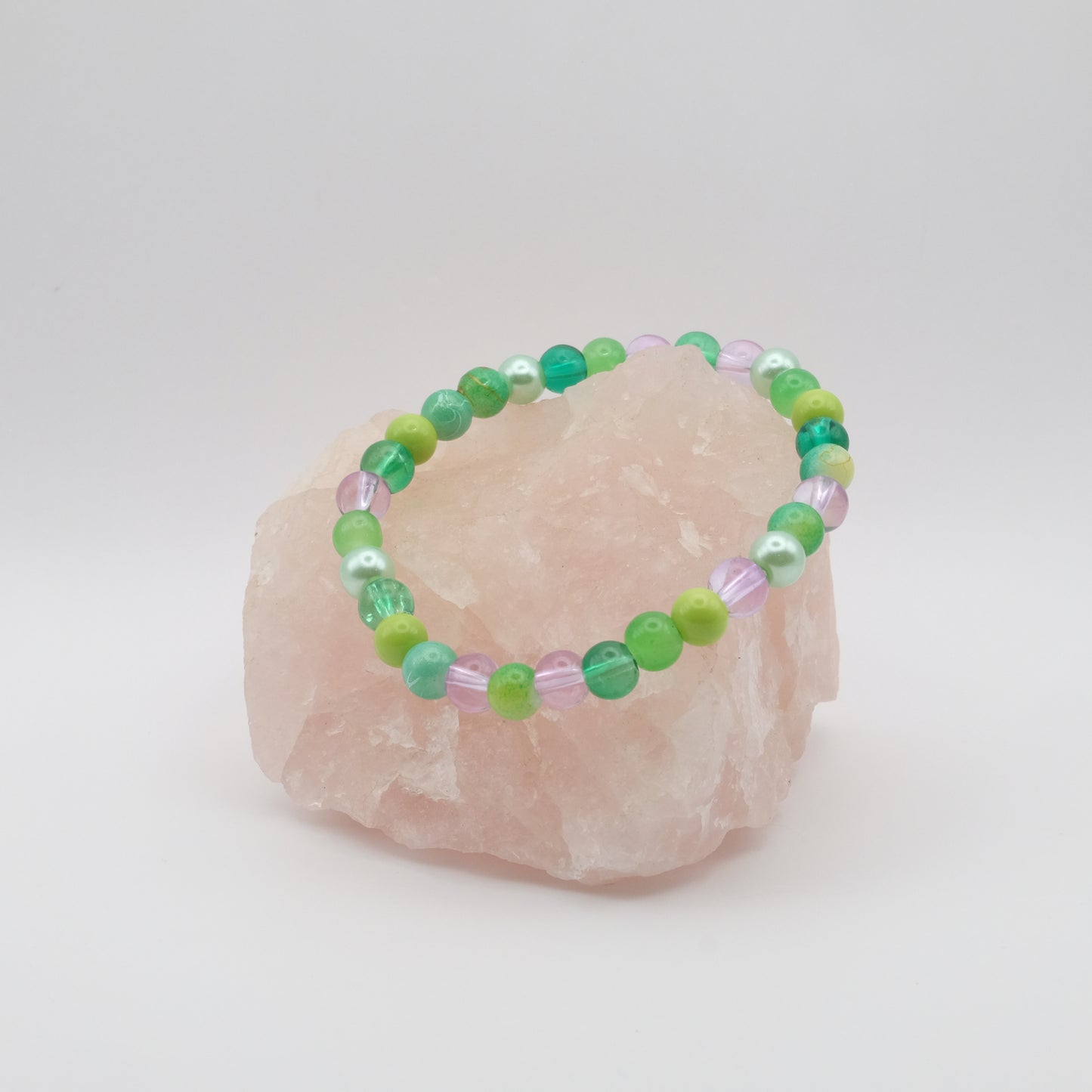 A purple, green and teal elastic beaded bracelet sitting on a rose quartz rock with a white background